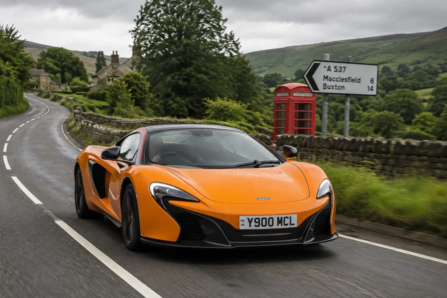 An orange McLaren sports car drives along a winding British country road on an overcast day. The wet tarmac reflects the car as it passes traditional stone walls, rolling green hills, a red telephone box and a UK road sign, capturing the scenic countryside and high-performance driving experience.