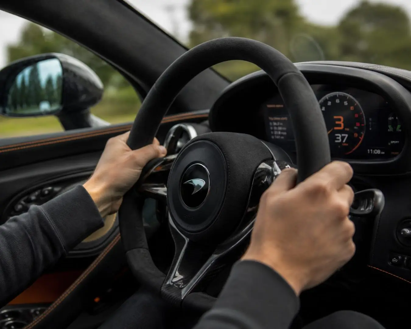 Close-up side-angle view of a driver's hands gripping the steering wheel inside a McLaren sports car. The premium leather and carbon fibre interior, digital instrument display and blurred scenery outside create a sense of speed, while the focus remains on the driver's hands and the high-performance cockpit.