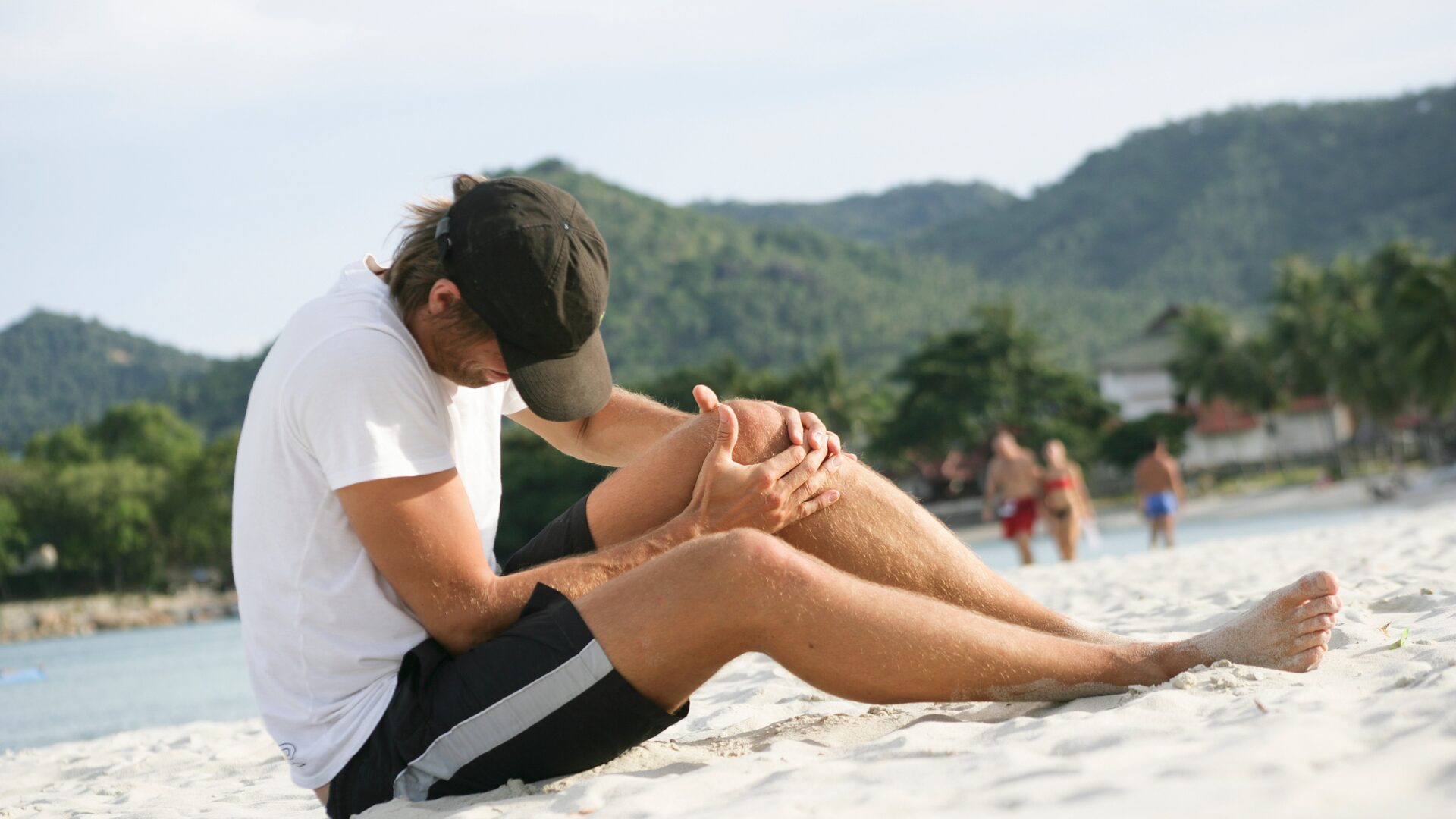 A man holding his knee in pain on a beach abroad.