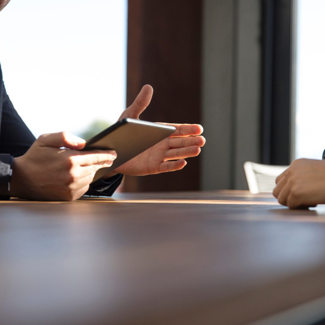 Two people sitting at a table in a modern office, discussing something on a digital tablet. One person gestures with their hand while holding the device.